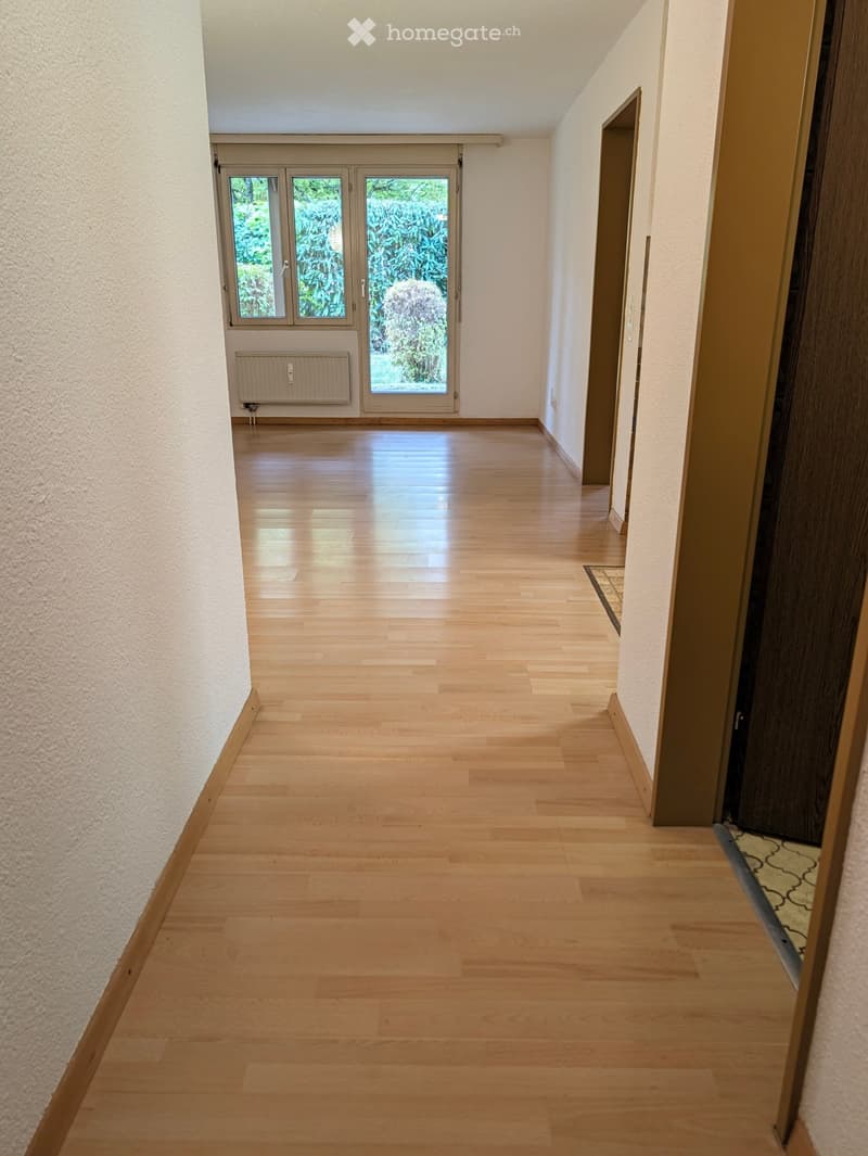 Empty hallway with light wooden flooring, white walls, and a large window at the end showing greenery outside.