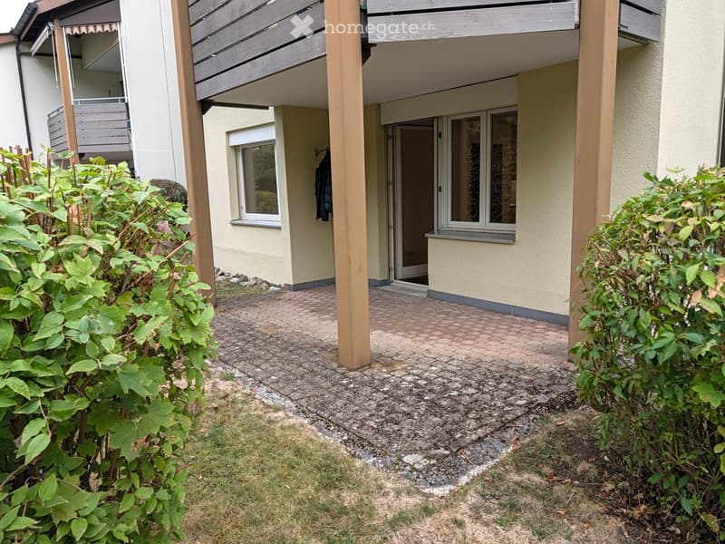 A small garden area with a paved pathway leading to a house entrance, surrounded by greenery and a wooden deck area above.