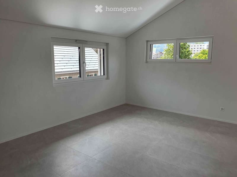 Empty room with light blue walls, tiled floor, and two windows offering views of the outside.