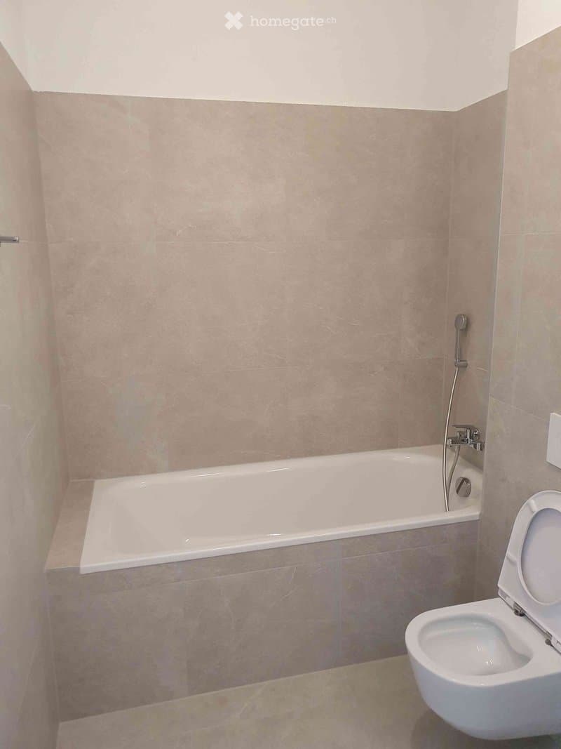 Modern bathroom with a bathtub, toilet, and beige tiled walls and floor.