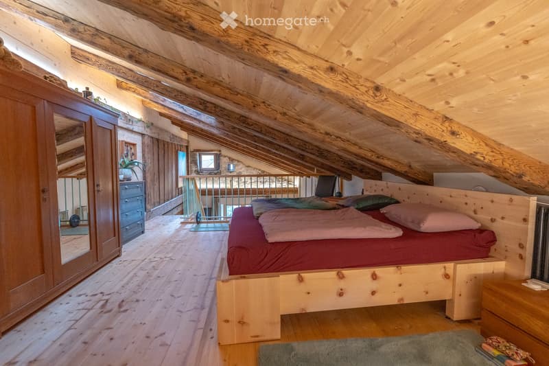Cozy attic bedroom with wooden beams, exposed wooden ceiling, and wooden floor. Features a wooden bed with red bedding, a large wardrobe with mirrored doors, a nightstand, and a small balcony area visible in the background.