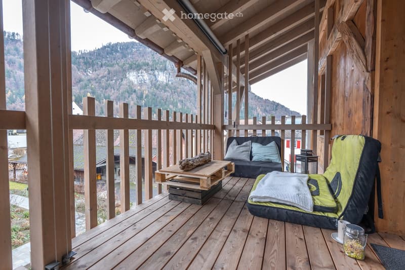 Wooden balcony with a scenic mountain view, featuring a sofa, small table with a log, backpack, and a cup on a surface.