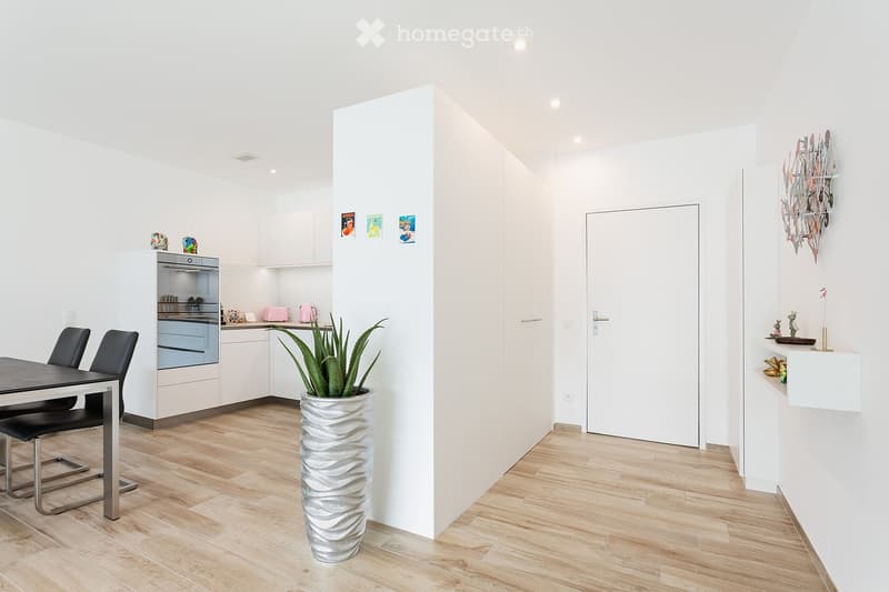 Modern, minimalist kitchen and dining area with white cabinets, a dining table with chairs, and a decorative plant in a vase.