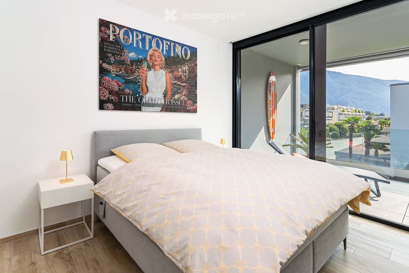 Modern bedroom with a large bed, nightstand, lamp, and a stylish poster on the wall. Large windows offer a scenic view of the landscape.