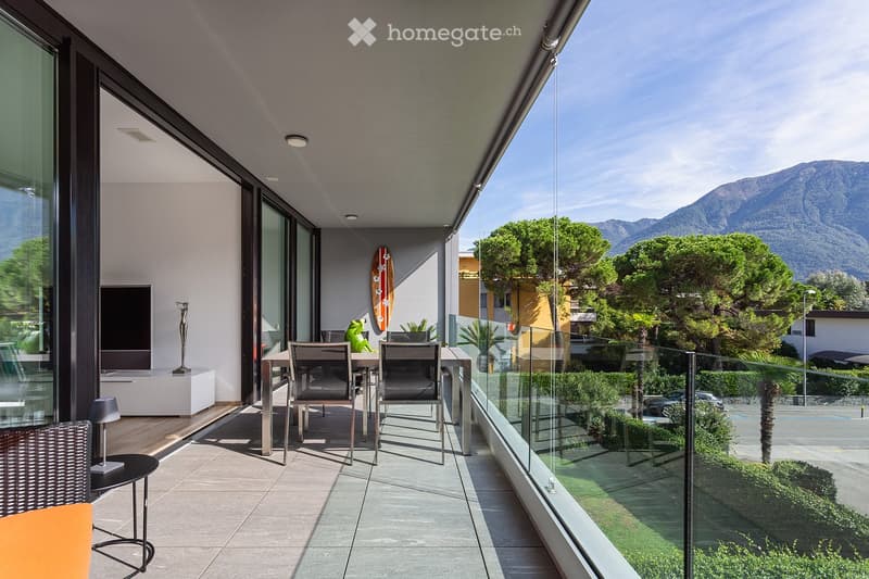 Modern balcony with glass railing, tiled floor, outdoor furniture including a table, chairs, and a stool, with a scenic mountain view in the background.