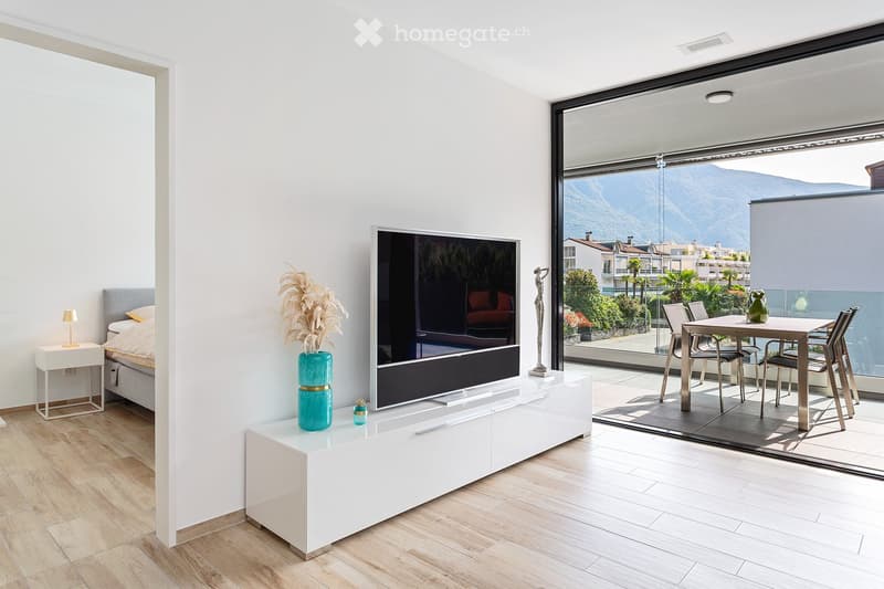 Modern living room with large windows offering a scenic mountain view, featuring a sofa, television, dining table, chairs, and decorative vases.