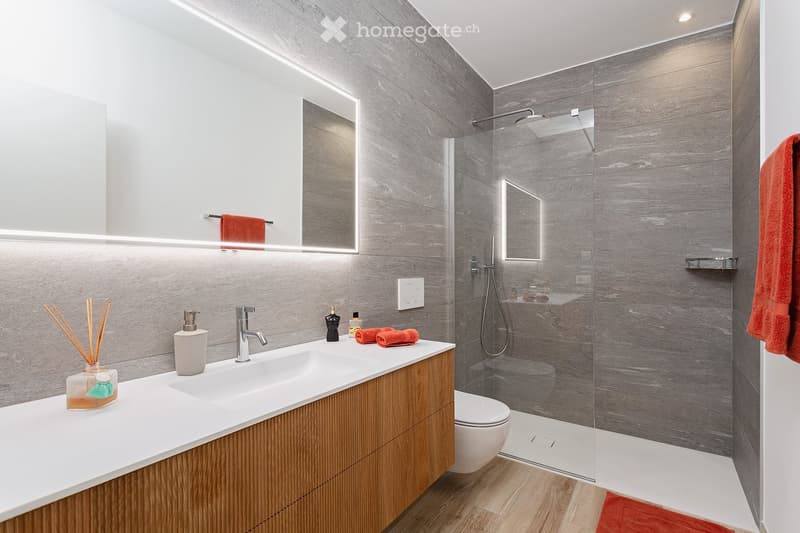 Modern bathroom with a double sink, sleek wooden vanity, grey tiled walls, a glass-enclosed shower, and a toilet. Features include a large mirror with built-in lighting, red towels, and decorative items on the sink.