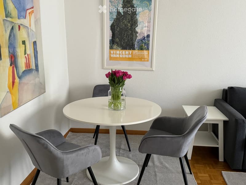 Modern dining area with a round white table, two grey chairs, a grey sofa, and a vase with pink tulips. Artwork and a poster are displayed on the wall.