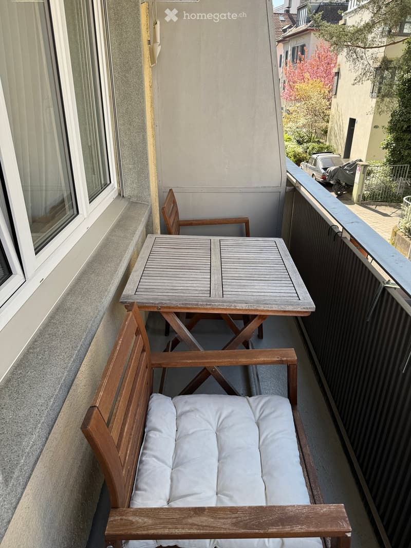Balcony with a wooden table, two chairs, and a bench with white cushions, overlooking a street with parked cars and trees.