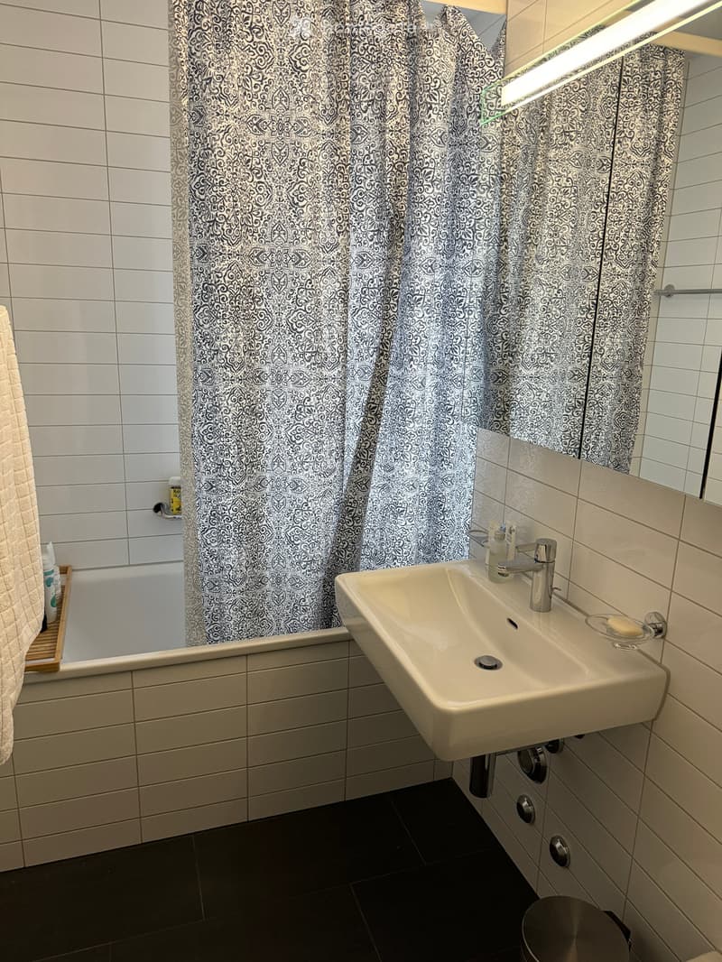 Modern bathroom with white tiles, a sink with a chrome faucet, a bathtub with a patterned shower curtain, a mirror, and a towel on a wooden rack.