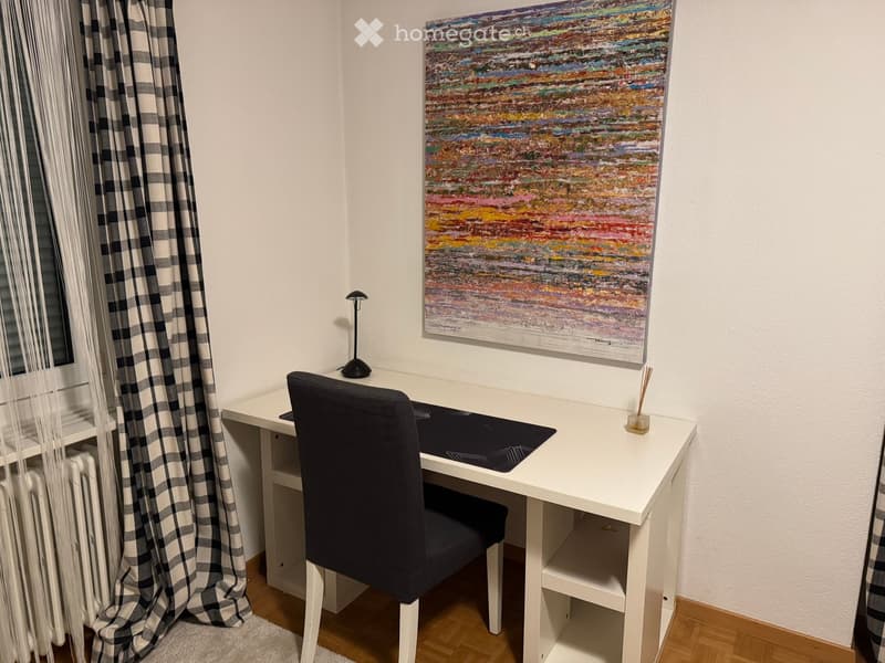 A small, well-lit room with a white desk, black chair, a colorful abstract painting on the wall, and a checkered curtain by the window.