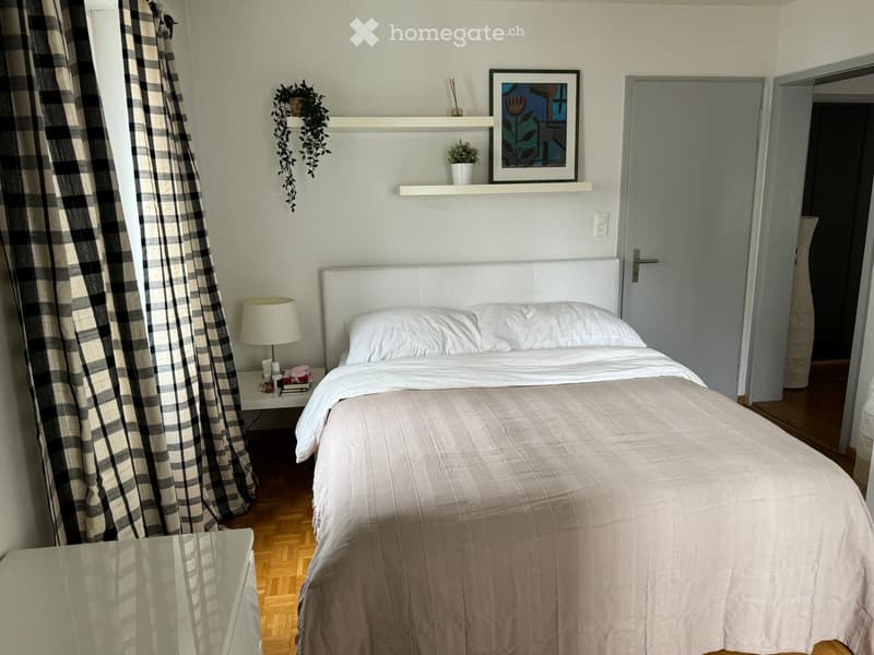 Cozy bedroom with a neatly made bed, nightstand with lamp, shelf with decorative items, and a window with plaid curtains.