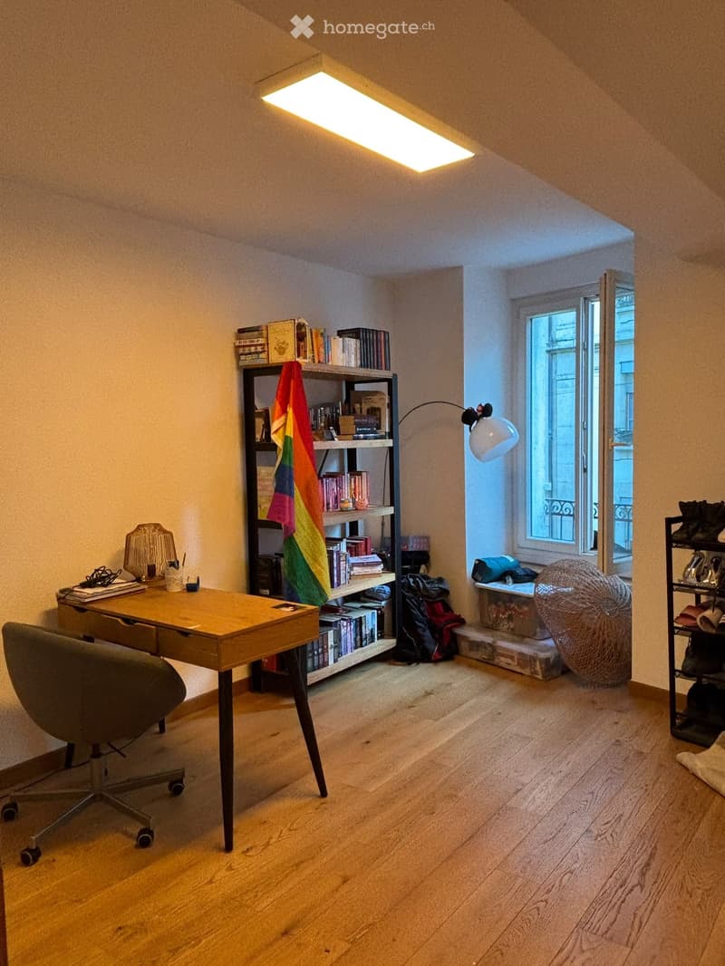A cozy, well-lit room with wooden flooring, a desk with a chair, a bookshelf filled with books and a colorful flag, a wicker ball, and a shoe rack near the window.