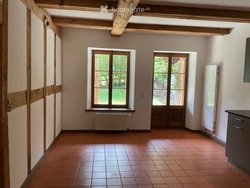 Empty room with wooden beams on the ceiling, tiled floor, large window, and glass door leading outside.