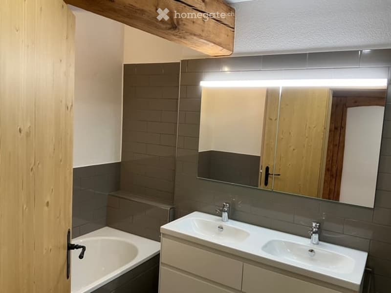 Modern bathroom with a bathtub, double sink, and large mirror with built-in lighting. Grey tiles and wooden accents.