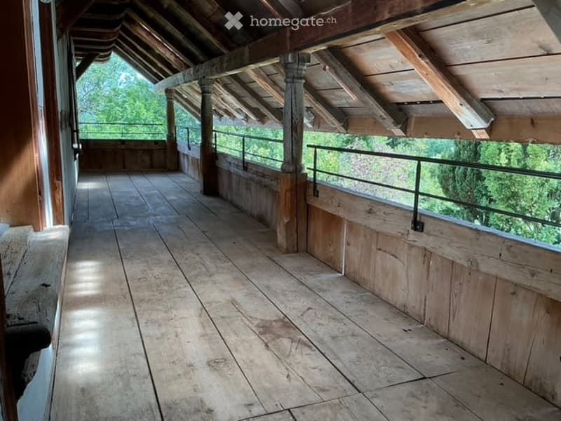 Wooden balcony with exposed beams, metal railing, and wooden flooring, overlooking a green outdoor area.