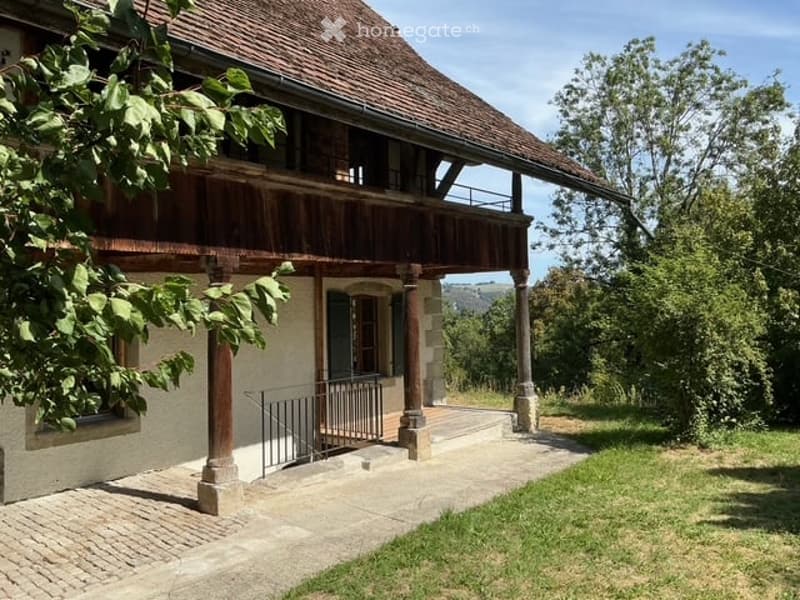 Two-story house with a wooden balcony, tiled roof, and stone pillars. The entrance features a metal gate and a paved pathway leading to the house.