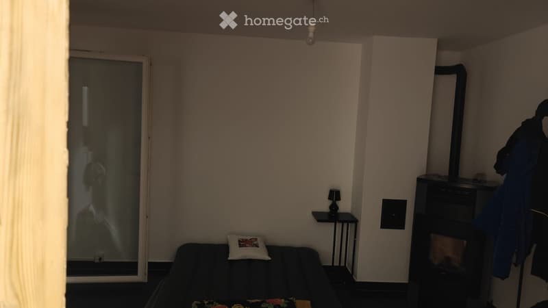 A small, modern living room with a dark-colored sofa, a decorative pillow, a small side table with a lamp, and a coat rack with jackets.