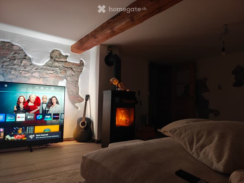 Cozy living room with exposed wooden beams, a stone wall, a fireplace, a television, a guitar, and a sofa.