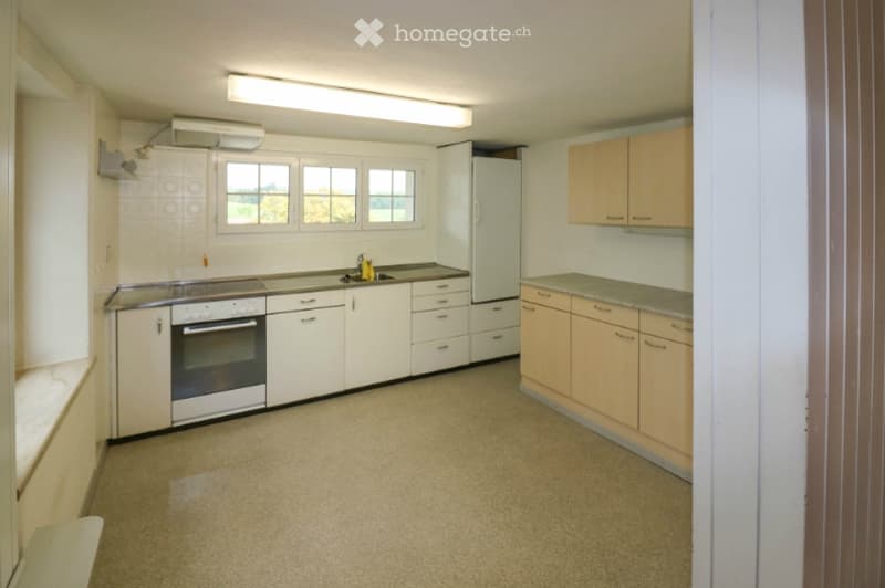 A small, unfurnished kitchen with white cabinets, a stainless steel oven, a refrigerator, and a sink. The space has a tiled backsplash and a window above the sink.