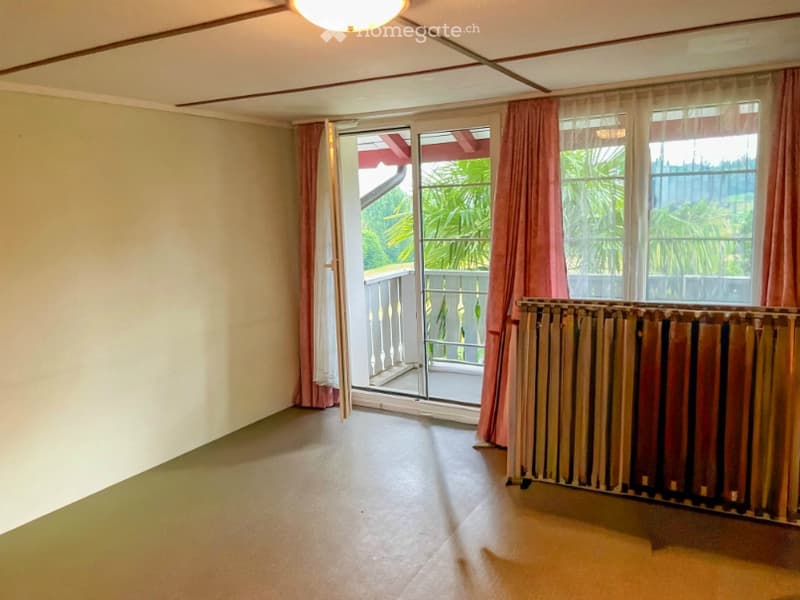 Empty room with light-colored walls, tiled floor, and a radiator. A sliding glass door opens to a balcony with a metal railing and outdoor plants visible.