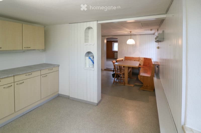 The image shows a small kitchen area with light-colored cabinets and a countertop on the left, connected to a dining area with a wooden table, chairs, and a bench. The walls are white, and the floor is carpeted.