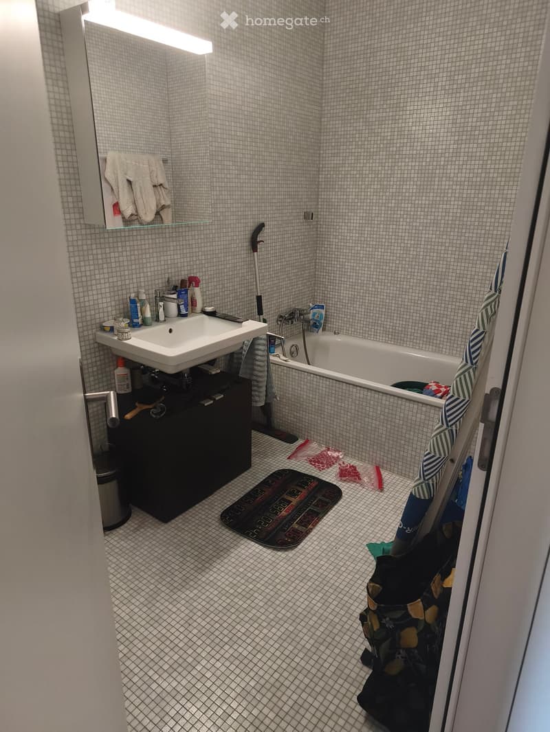 Small bathroom with white tiled walls and floor, featuring a sink with a cabinet underneath, a bathtub, a mirror with a towel hanging on it, and a bathmat on the floor.