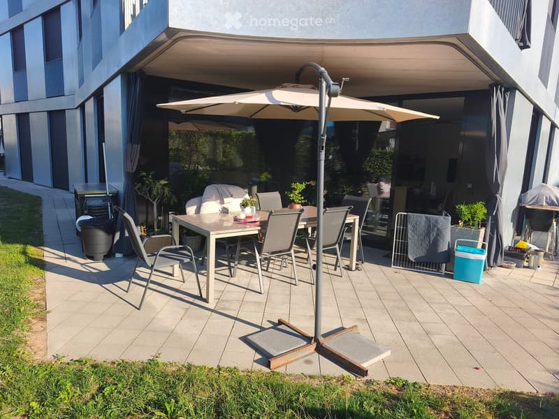A modern outdoor patio area with a large umbrella, a dining table with chairs, potted plants, and a grill. The patio is connected to a building with large glass doors and curtains.