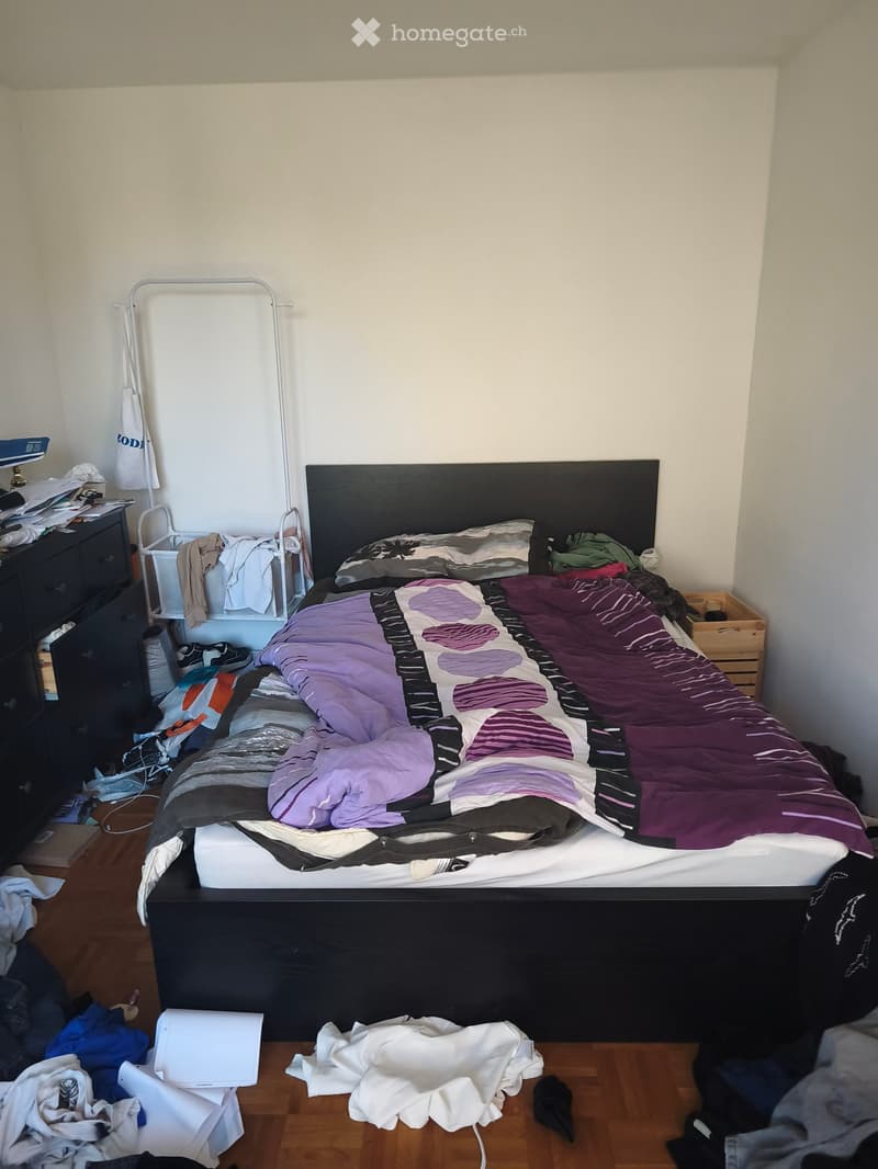 A bedroom with a neatly made bed, a bedside table, a wardrobe, and a clothing rack. The room has wooden flooring and white walls.