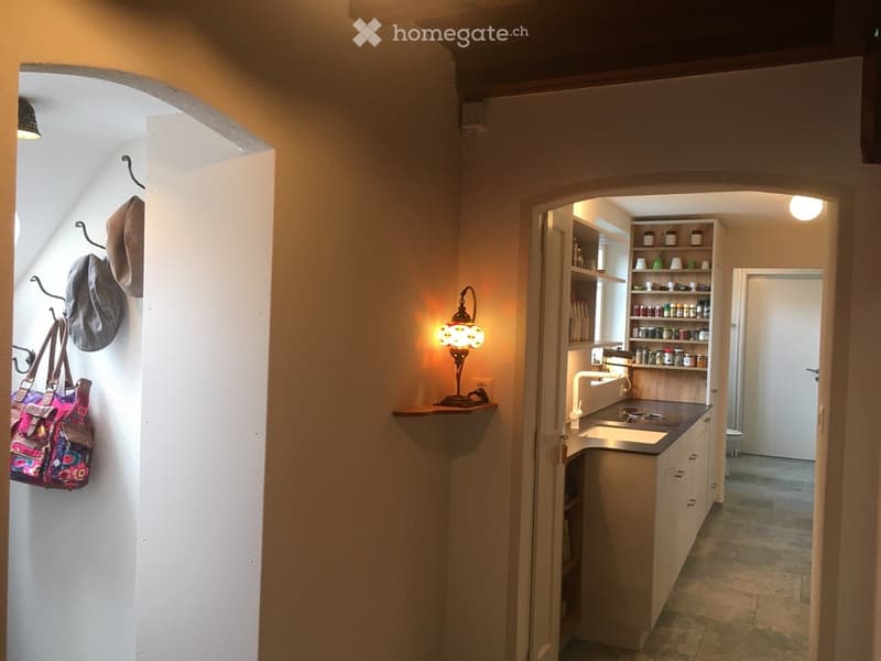 A narrow hallway with slanted ceilings, leading to a small kitchen area. The hallway features a coat rack with hats and a bag, and the kitchen has a sink, countertop, and shelves with jars and bottles.