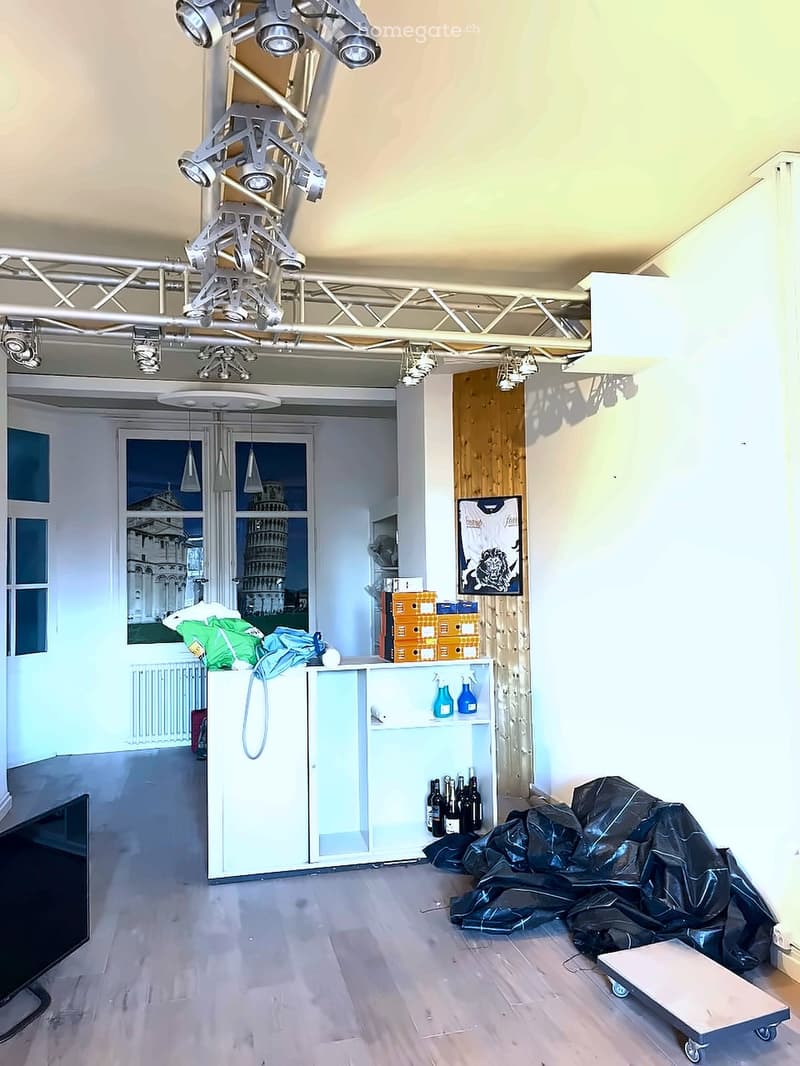 The room features a modern, industrial design with exposed lighting rigs, a large window showing a scenic view, a cabinet with bottles, and a covered object on the floor.