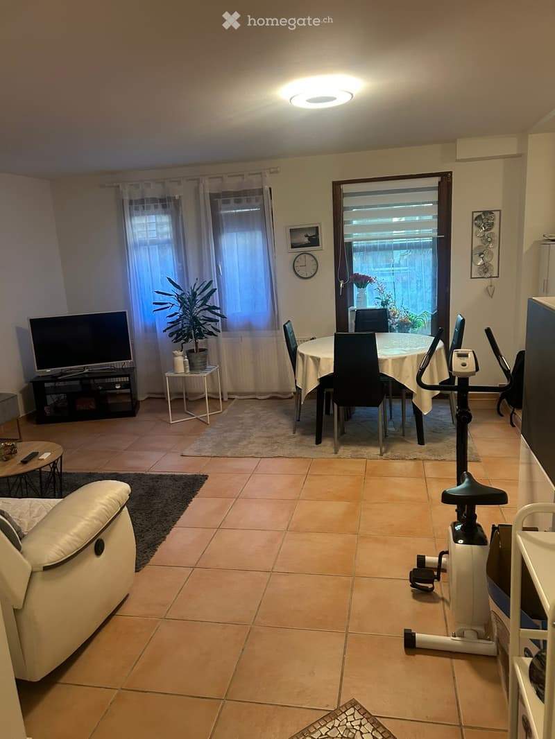A well-lit living and dining area with a cream sofa, dining table with chairs, TV, exercise bike, and decorative plant. The space features tiled flooring and large windows with sheer curtains.