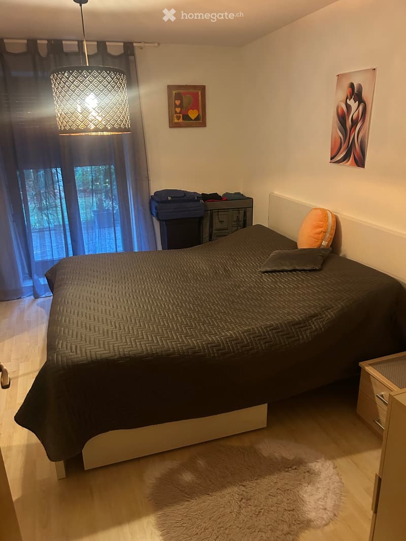 A modern bedroom with a large bed, dark bedding, a nightstand, a hanging lamp, and a large window with dark curtains.