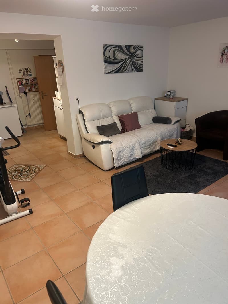 Living room with a white sofa, small round table, and a chair. The room features tiled flooring and a doorway leading to another room.