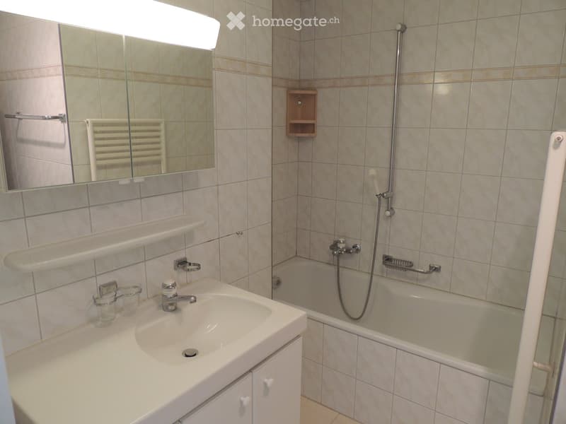 Bathroom with a white sink, bathtub, mirror, and wooden shelf. The walls are tiled with beige tiles and brown accents.