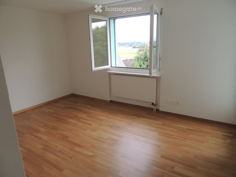 Empty room with wooden flooring, white walls, and large windows offering a view of greenery and a landscape.