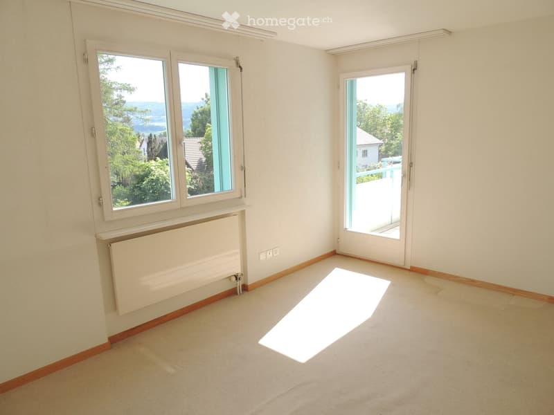 Empty room with light-colored walls, wooden baseboards, carpeted floor, large windows, and a sliding glass door leading to a balcony.