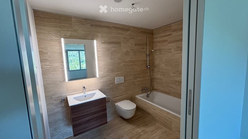 Modern bathroom with wooden wall tiles, a white sink, toilet, bathtub, and a large mirror with built-in lighting. The space features a sleek, contemporary design with a showerhead visible above the bathtub.