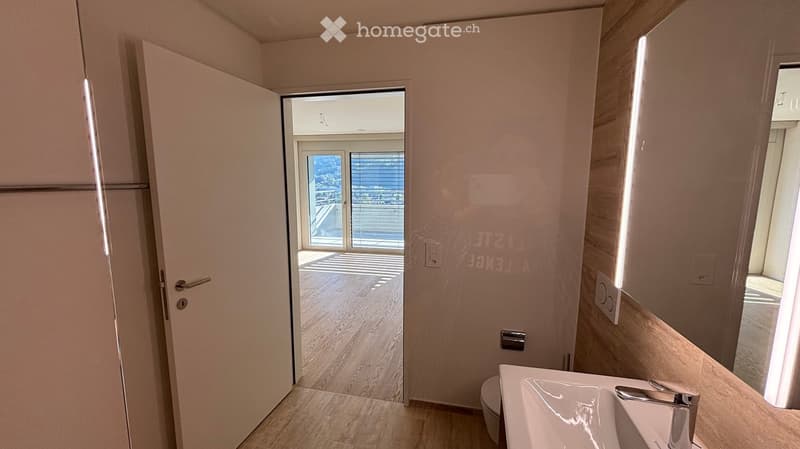 Modern bathroom with a white sink, chrome faucet, and toilet. The room features light-colored tiles and a wooden floor, with a door leading to a balcony or outdoor area.