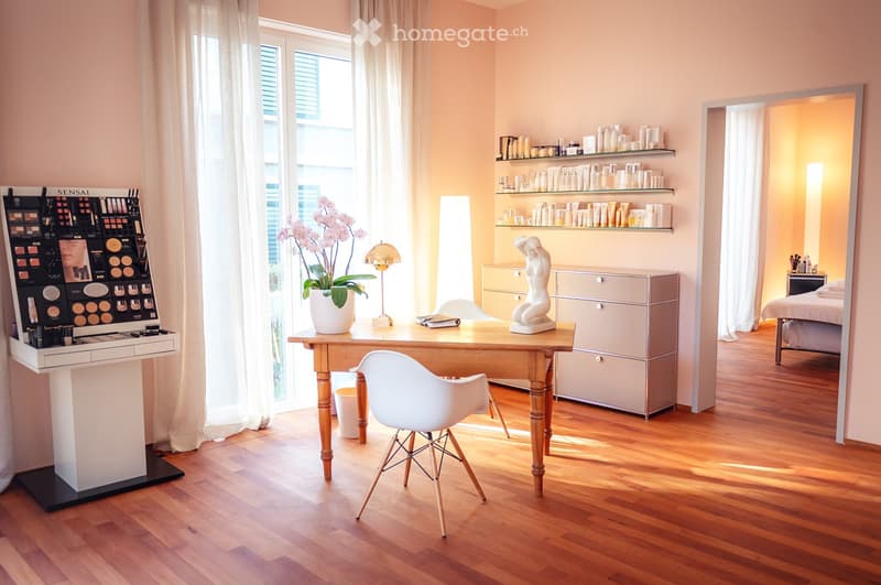 Furnished Room. Well-lit room with wooden flooring, large window with sheer curtains, makeup display, wooden table with a lamp, vase with flowers, white chair, shelves with various items, and a bed visible in the adjacent room.