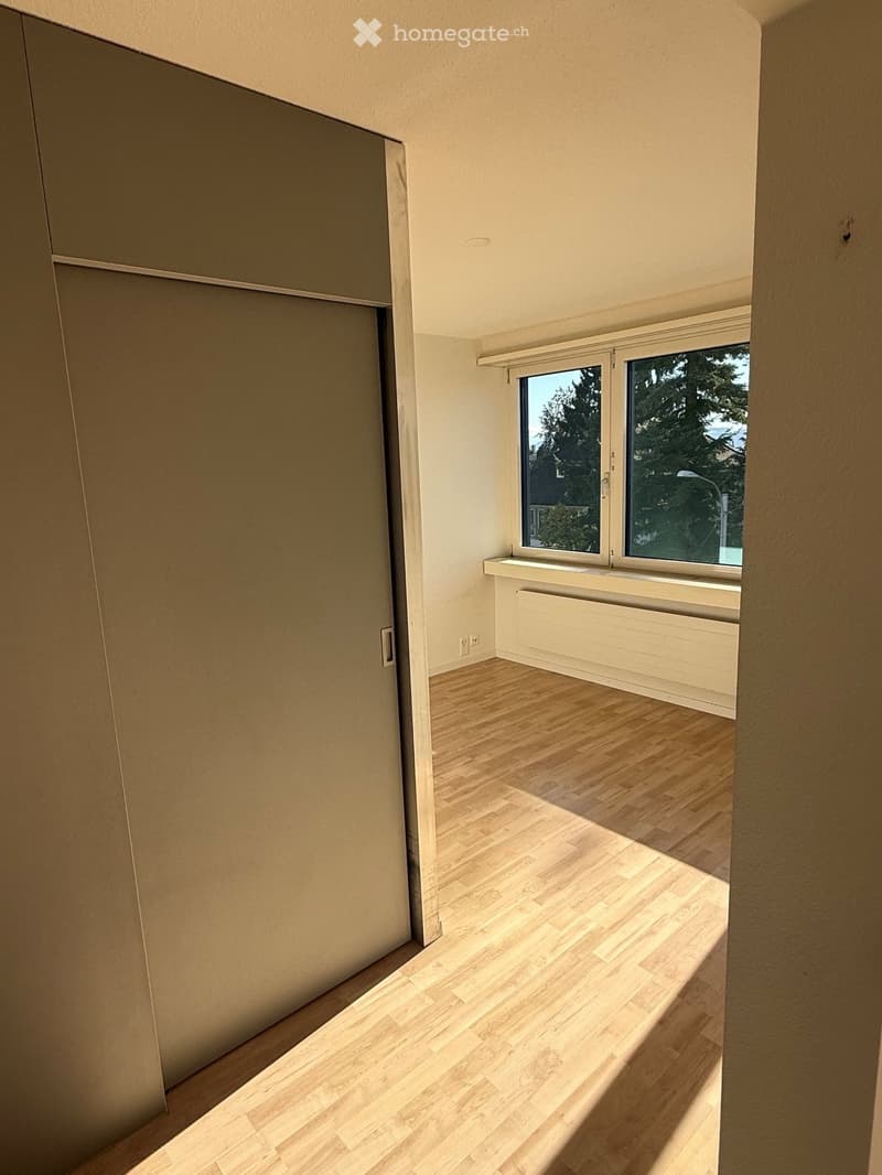 Empty room with light wooden flooring, white walls, and a large window with a view of trees. No furniture or equipment is present.
