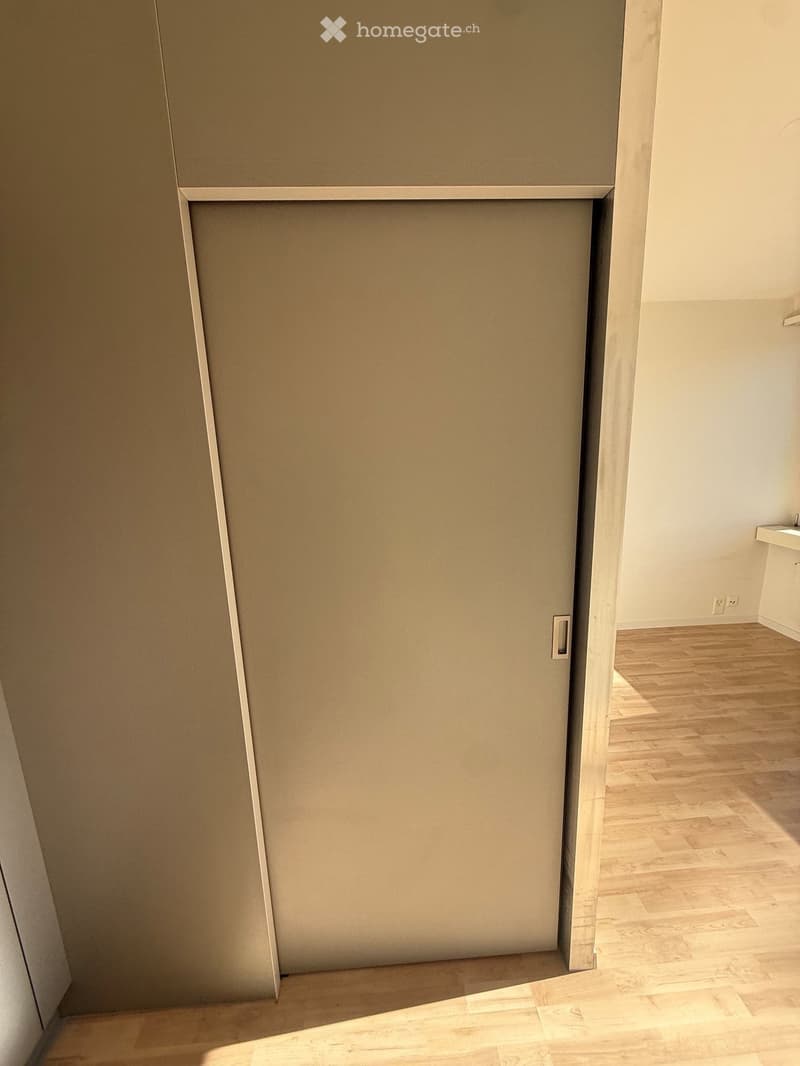 Empty room with wooden flooring and a closed door leading to another room. The walls are painted white, and the space is well-lit.