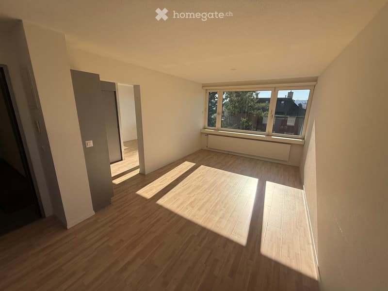 Empty room with wooden flooring, white walls, and large windows allowing natural light to enter. The room features a radiator below the windows and an open doorway leading to another space.