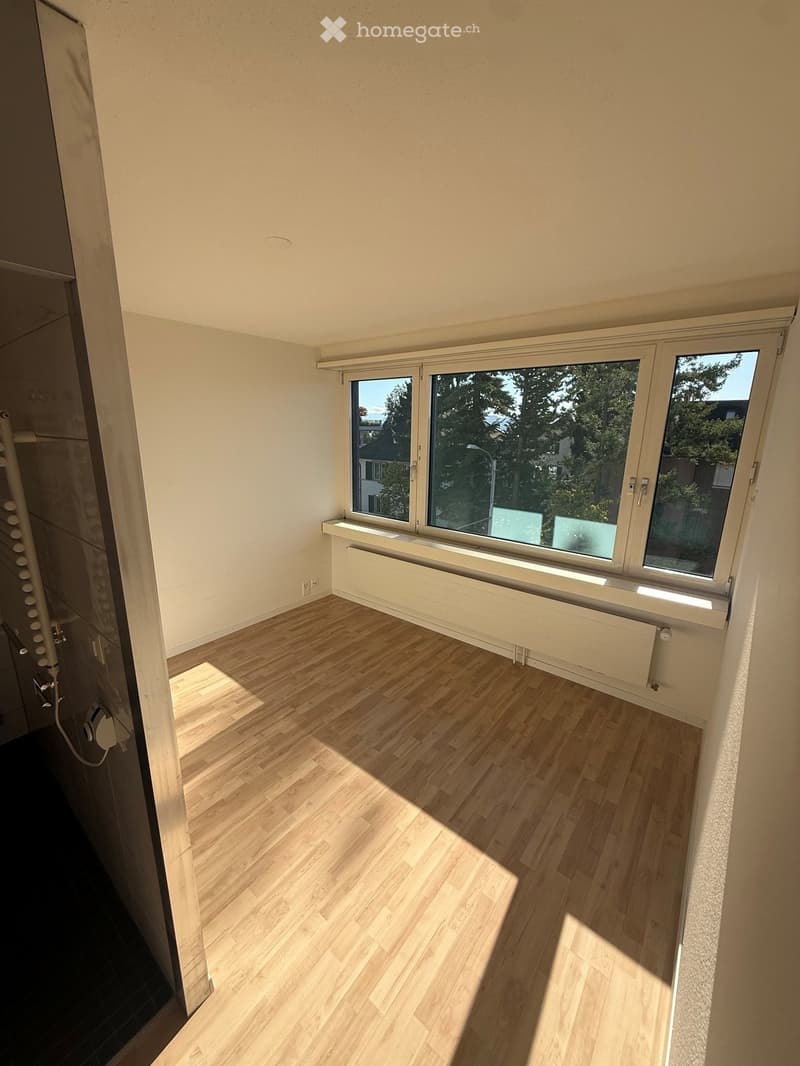Empty room with light wooden flooring, white walls, and large windows offering a view of trees and neighboring buildings.