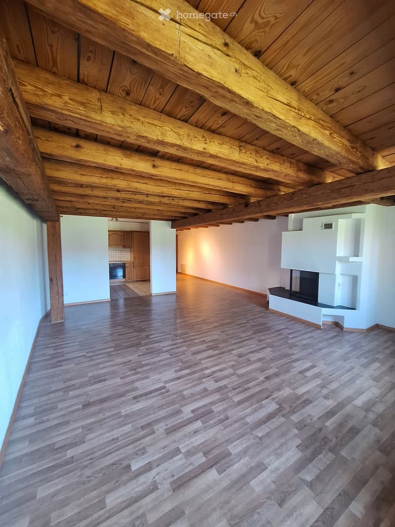 Empty room with wooden floor and exposed wooden beams on the ceiling. The room has white walls and a fireplace on the right side. The background shows a kitchen area with cabinets and an oven.