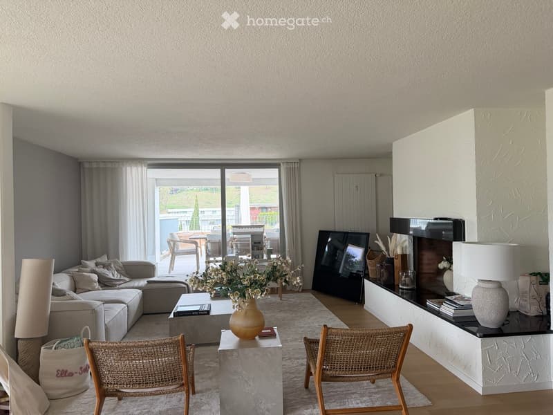 Modern living room with a grey sectional sofa, coffee table, wicker chairs, a large TV, and a fireplace with decorative items. Large windows offer a view of the outdoor area.