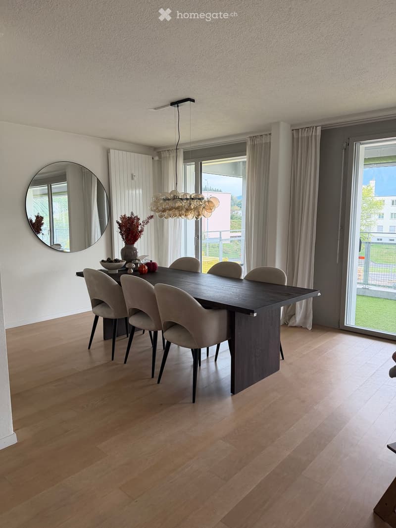Modern dining room with a dark wooden dining table, six beige chairs, a large round mirror, a decorative vase with dried flowers, and a modern chandelier.