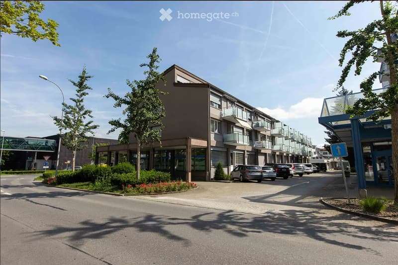 Modern multi-story apartment building with balconies, surrounded by trees and parked cars in a paved area.