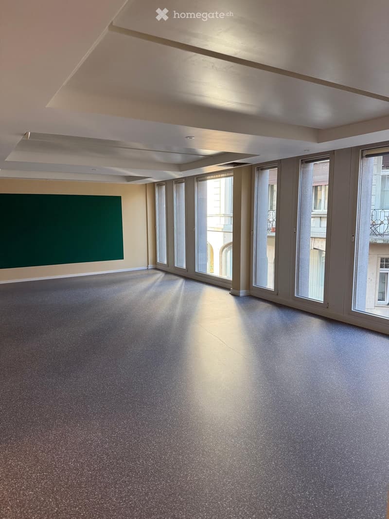 Empty room with grey carpeted floor, large windows, and a green wall panel.
