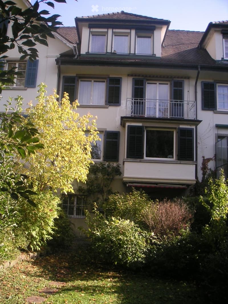 Two-story house with multiple windows, balconies, and surrounding greenery.